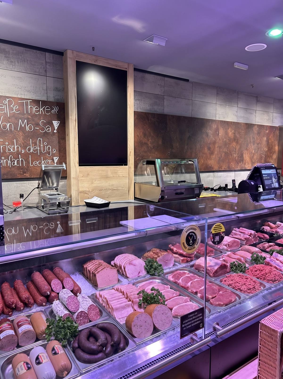Die Bedientheke des Marktes mit Wurst, Aufschnitt und frischem Fleisch auf Edelstahlschalen, dahinter die Tafel mit den Öffnungszeiten der Theke.
