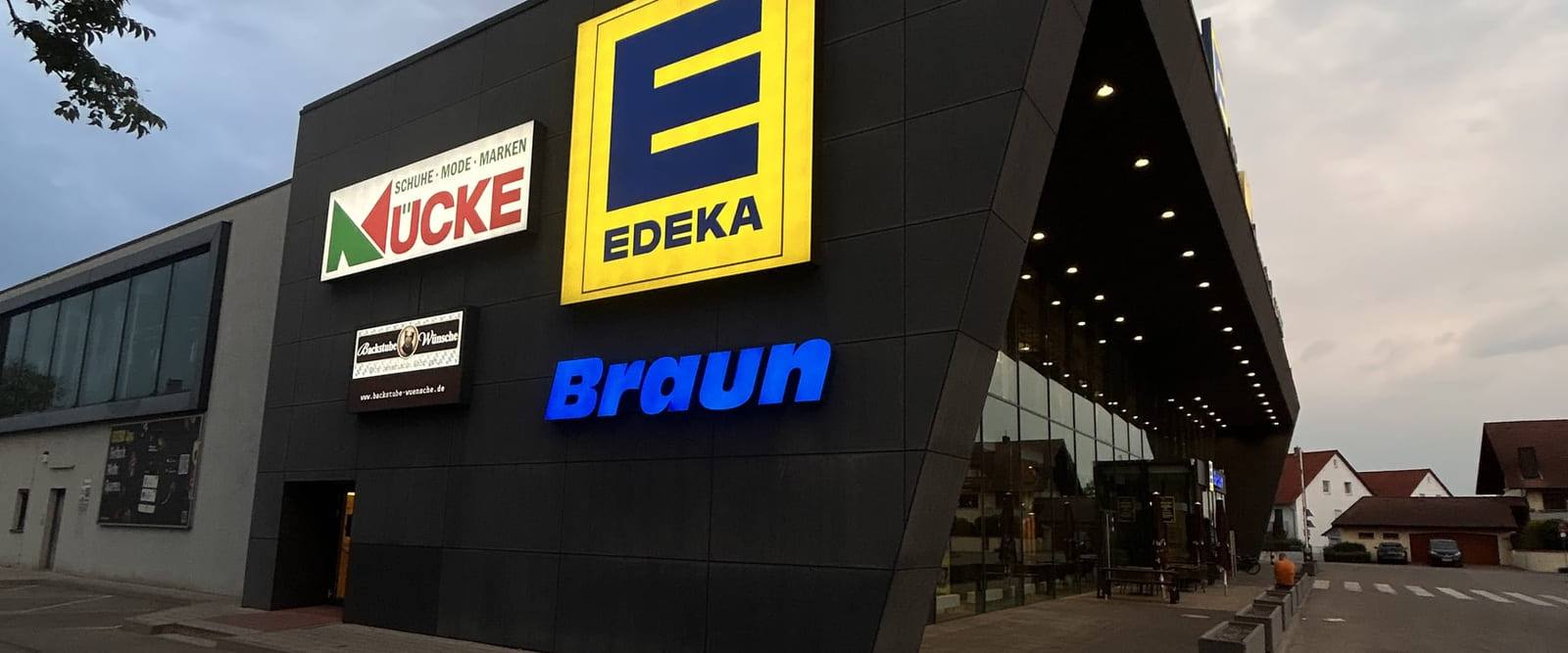 Das EDEKA Braun Center in der Dämmerung: die spitze Dachkante über dem beleuchteten Eingang, an der Fassade die Schilder von EDEKA Braun, MUCKE und Backstube Wünsche.