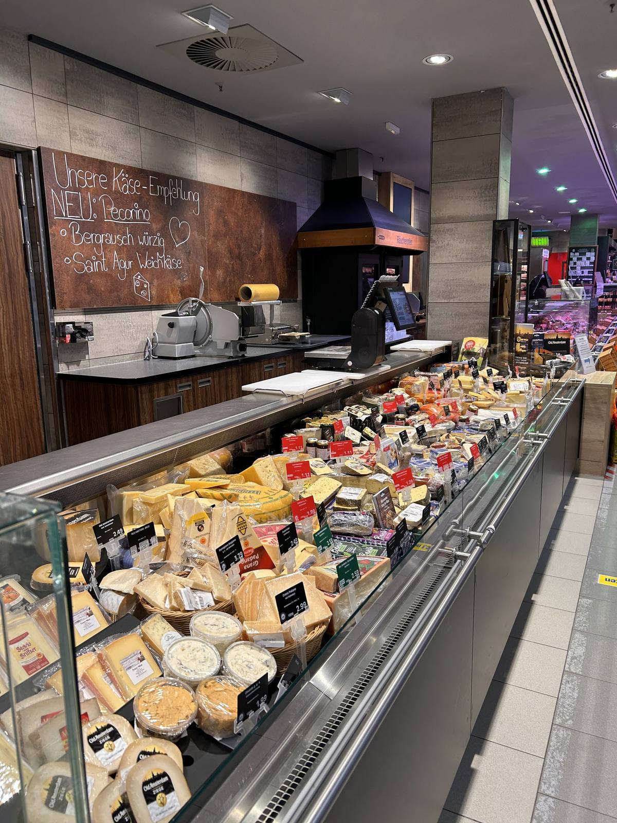 Die Käsetheke in ganzer Länge, dicht bestückt mit Laiben, Stücken und Frischkäse in Körben. An der Wand eine handgeschriebene Tafel mit der Käse-Empfehlung der Woche.