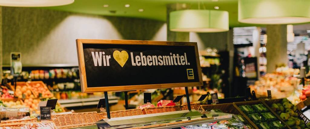 Das gerahmte Schild „Wir lieben Lebensmittel“ mit gelbem Herz über der Obstauslage, dahinter die grüne Decke der Markthalle.